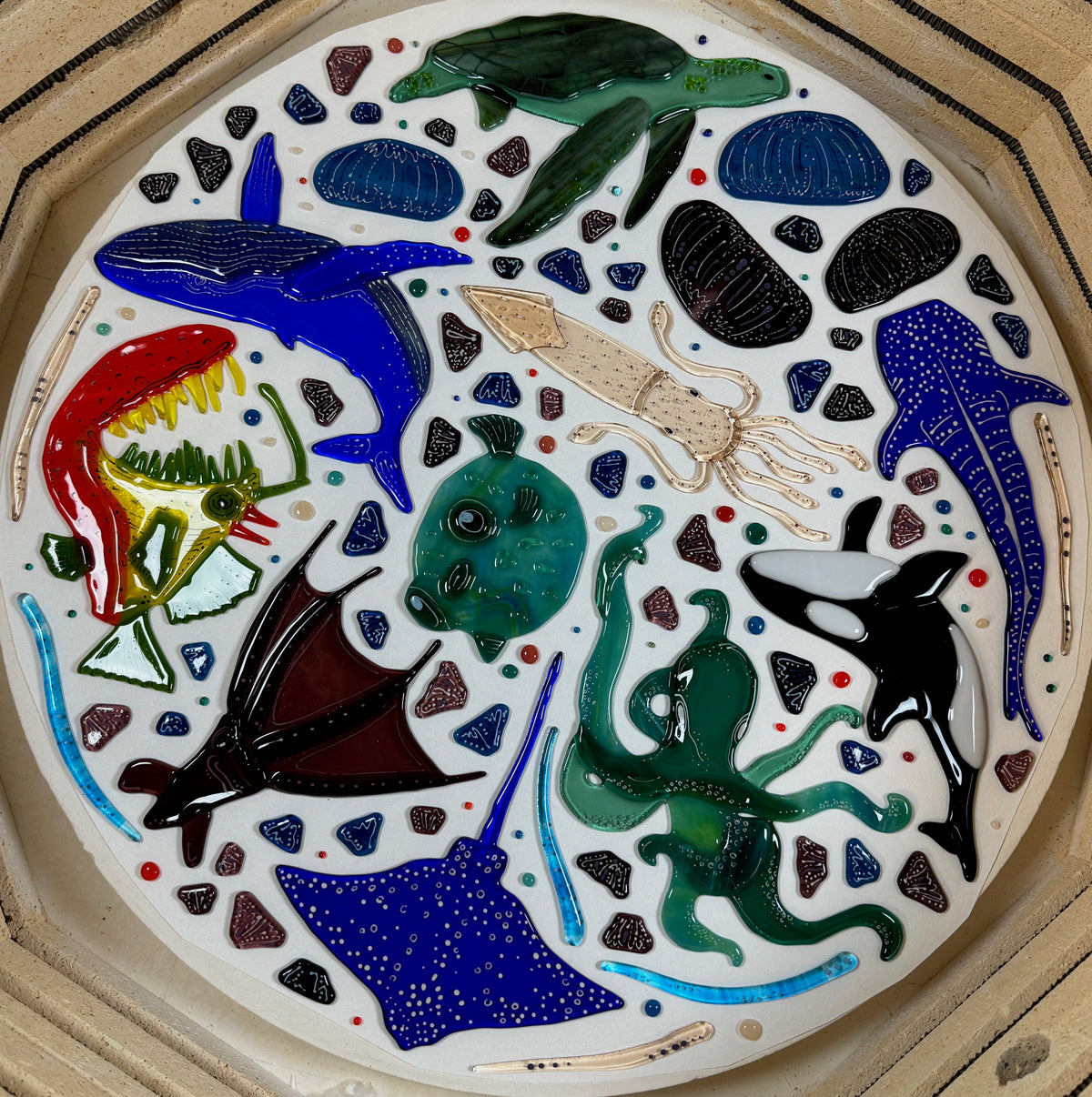 A white tray featuring colorful handmade fused glass art pieces with ocean creatures including whales, anglerfish, an octopus, a stingray, a turtle, and a squid, surrounded by small fish and shell-like motifs within a kiln. 
