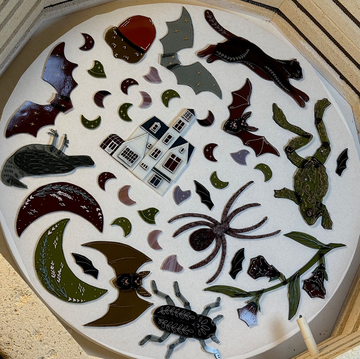 A circular white board or tray displaying a collection of handmade fused glass pieces, including a haunted house, spider, bat, frog, beetle, moon phases, crescent moons, and other witchy-themed motifs, arranged within a kiln. 